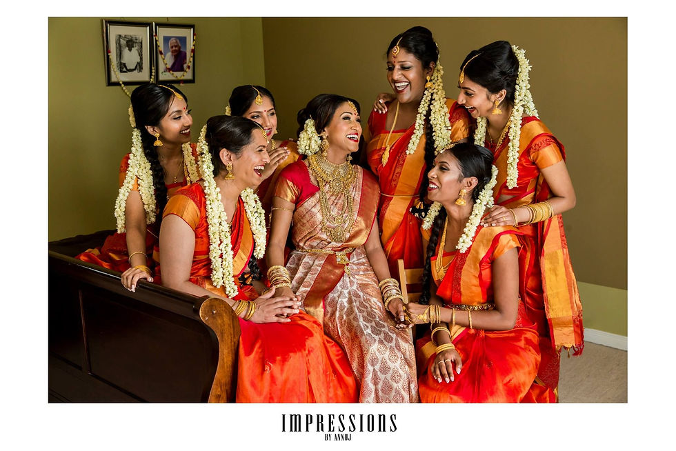 A joyful bride dressed in a traditional silk saree, adorned with gold jewelry, is surrounded by her bridesmaids in matching vibrant orange and red sarees. The bridesmaids, also wearing traditional jewelry and white jasmine garlands in their hair, are sharing a light-hearted moment with the bride, capturing the essence of Kannada bridal celebrations.
