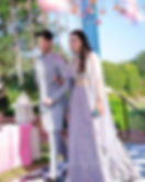 A couple dressed in coordinated pastel lavender outfits; the groom in an intricately designed sherwani and the bride in a mirror-work lehenga, posing under pastel floral decor at an outdoor wedding venue.