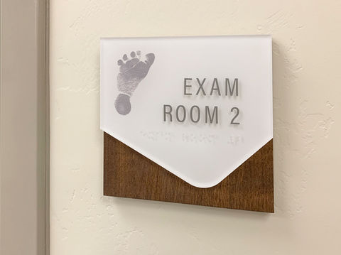Custom ADA room ID, "Exam Room 2" with a printed child's footprint design to the left. Frosted acrylic with raised lettering and braille, with a custom wood backer for dimension.