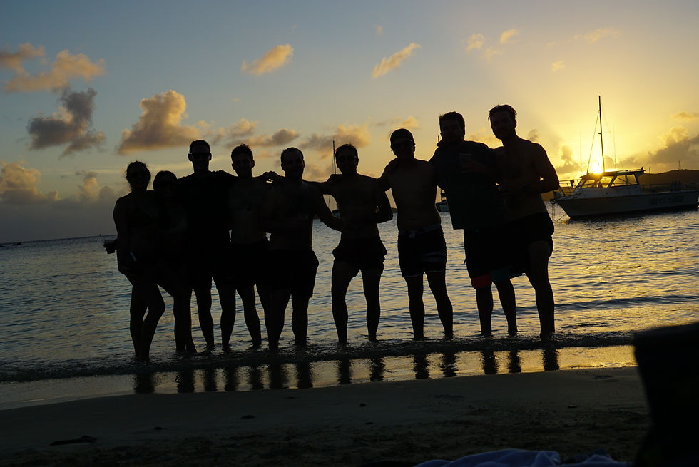 Silhouetted group of people stands on a beach at sunset, with boats in the background and a colorful sky, creating a joyful mood.