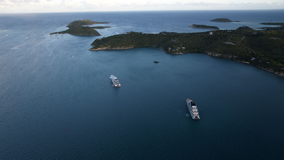 Aerial view of two yachts anchored in a calm, deep blue sea. Green islands dot the background under a partly cloudy sky.