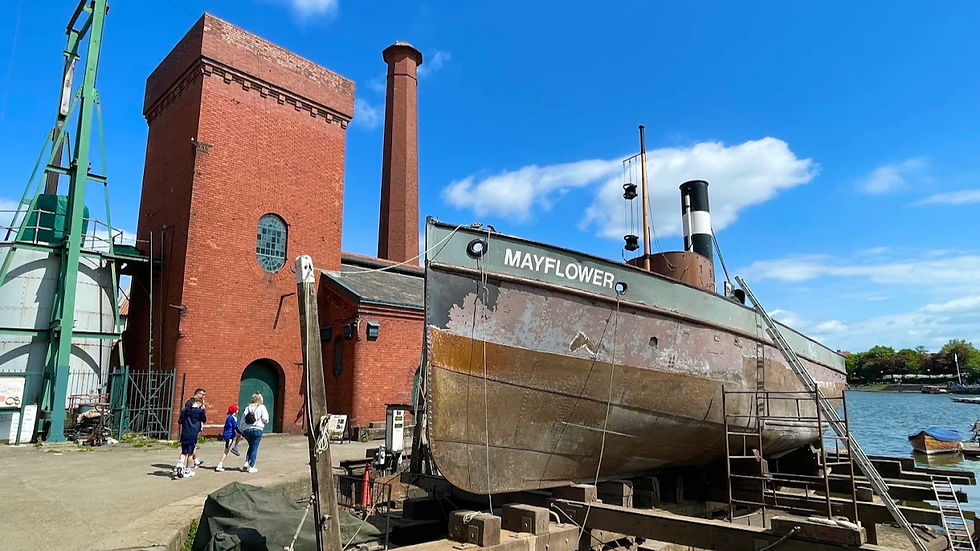 Historic boat Mayflower on dry dock beside red brick buildings, with visitors walking under a bright blue sky.