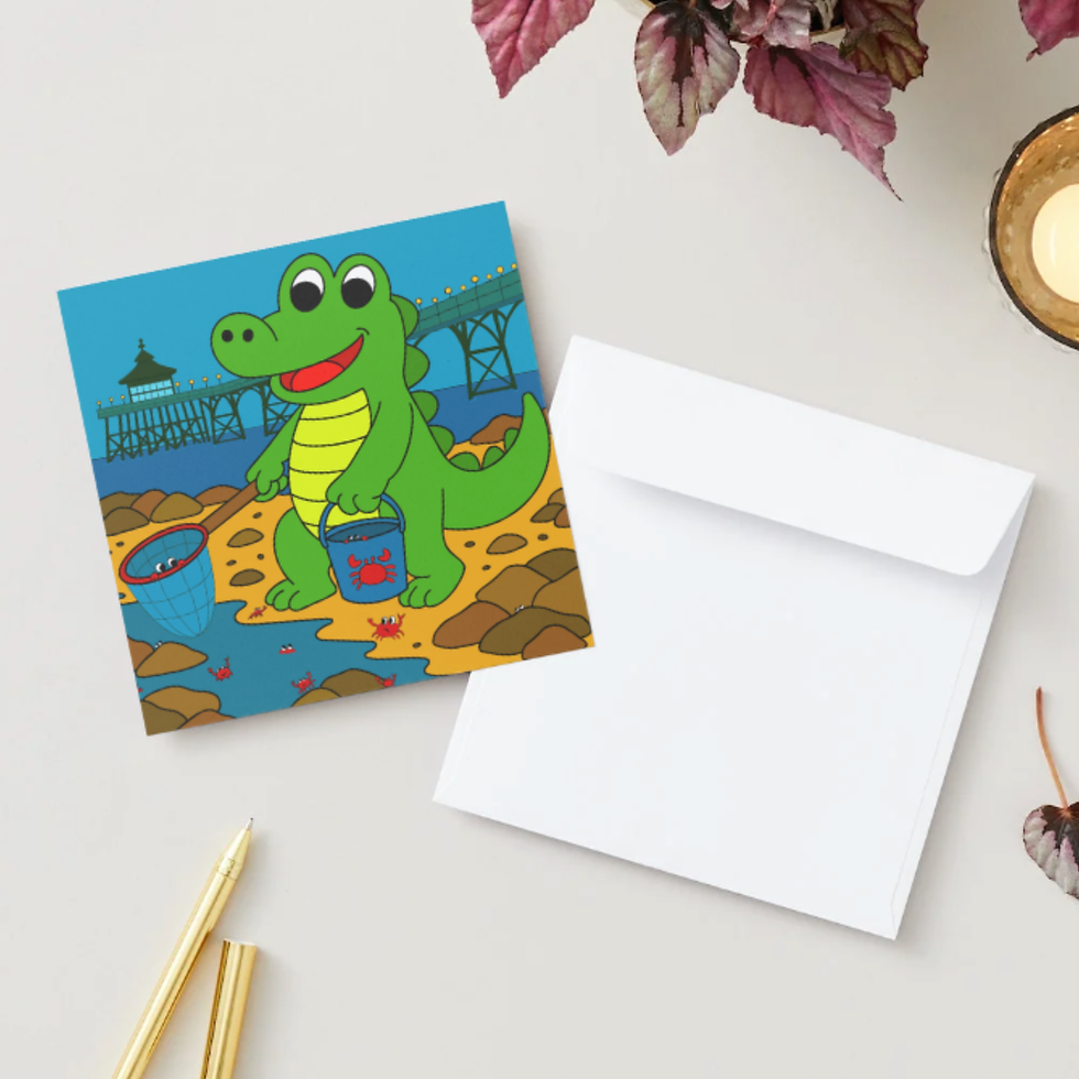 Square greeting card of crocodile crabbing by Clevedon Pier, beside white envelope, gold pens, candle, and leafy plant