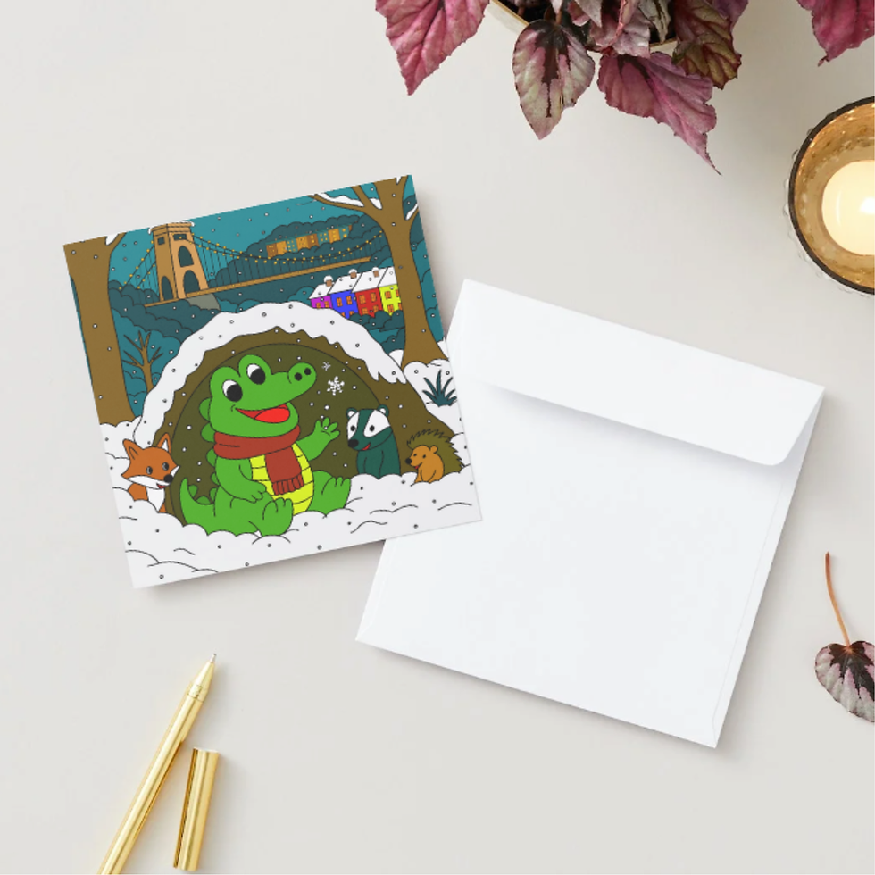 Square holiday card with crocodile at Clifton Suspension Bridge beside white envelope, gold pens, candle, and leafy plant