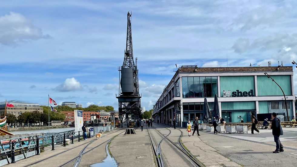 People walk along a waterfront pier beside a tall crane and the MShed building under a cloudy blue sky, with tracks in the pavement.