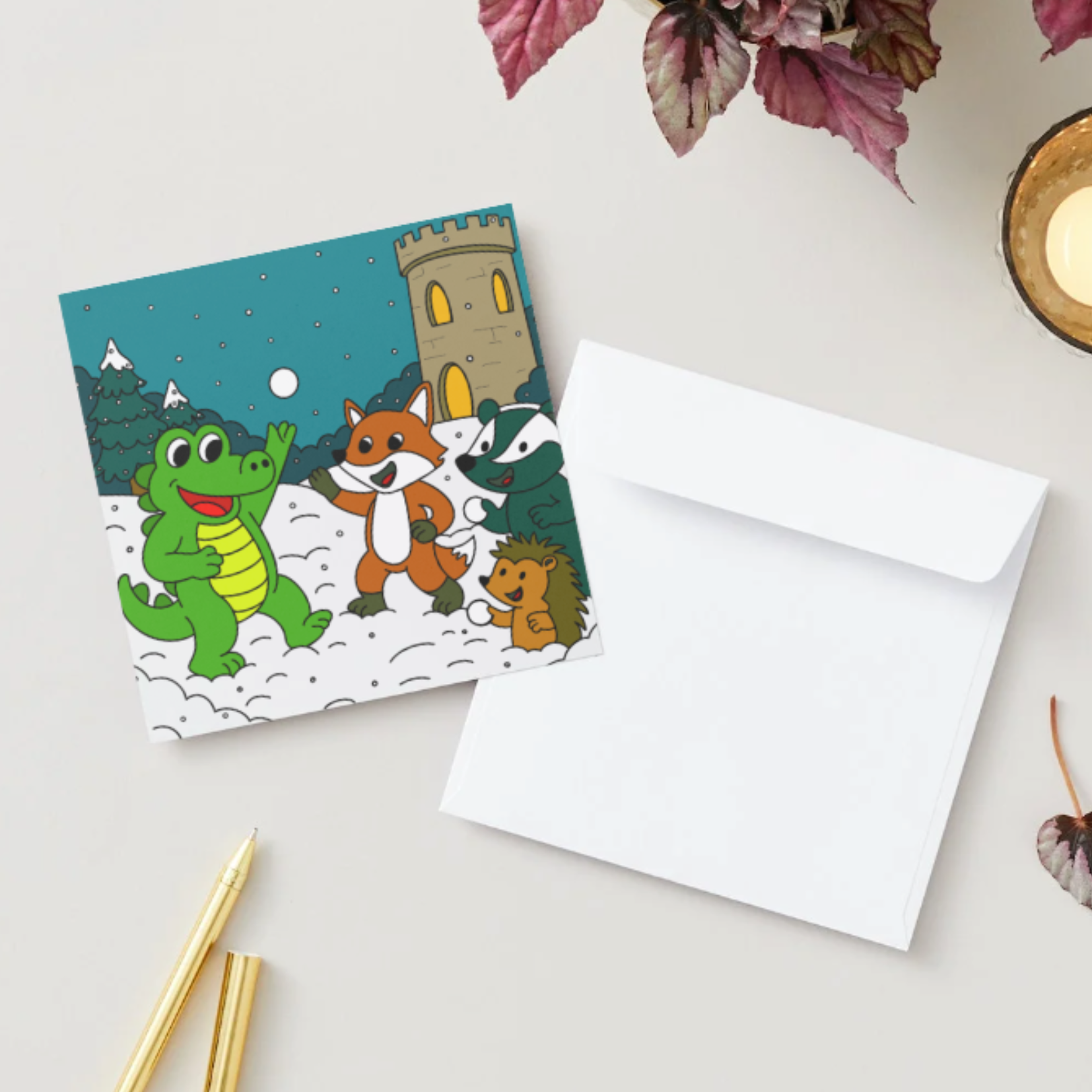 Square holiday card of crocodile in a snowball fight with friends, beside white envelope, gold pens, candle, and leafy plant