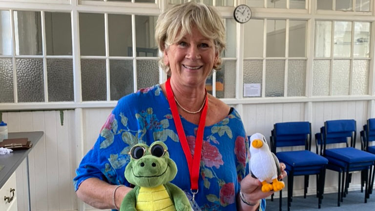 Author Kat Yeadon in blue floral top and red lanyard holds a plush crocodile and seagull in a bright room with blue chairs in the background.