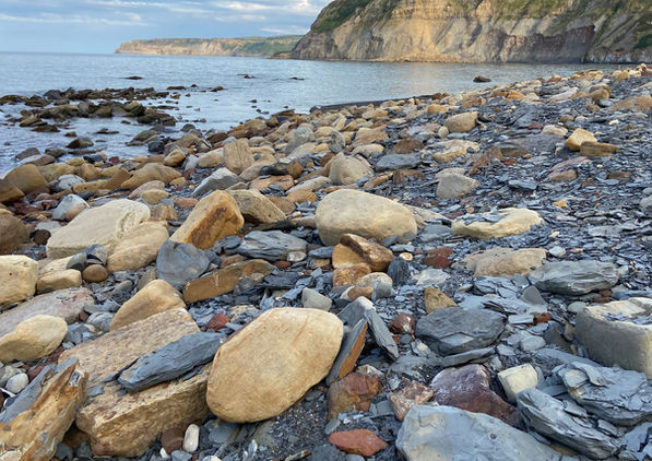 A rocky shoreline with large scattered stones, calm water, and steep sunlit cliffs beneath a partly cloudy sky.