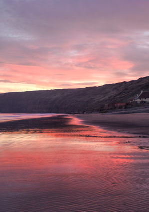 Saltburn beach at sunrise with pink and purple reflections on wet sand, cliffs in the distance, and houses along the shore.