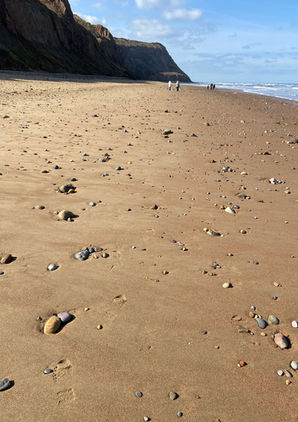 A wide sandy beach scattered with small stones, backed by tall cliffs under a bright blue sky.