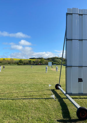 Cricket match in a grassy field near Loftus, with players in white and a large sight screen under a bright blue sky.