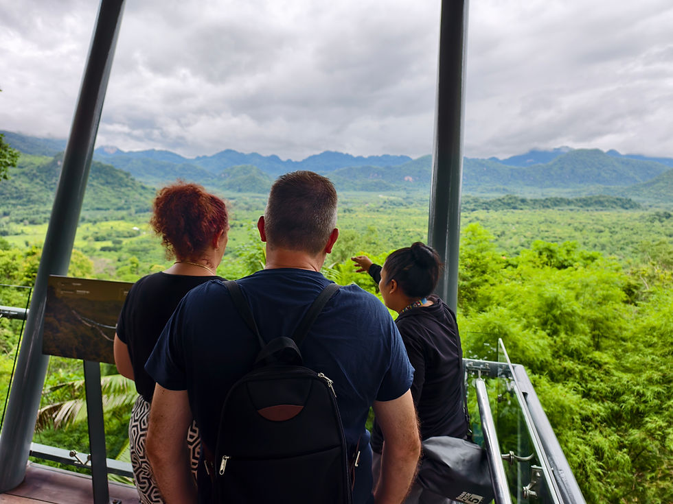 Three people stand on a viewing platform overlooking a lush green valley with mountains. One person points, possibly giving a tour. Cloudy sky.