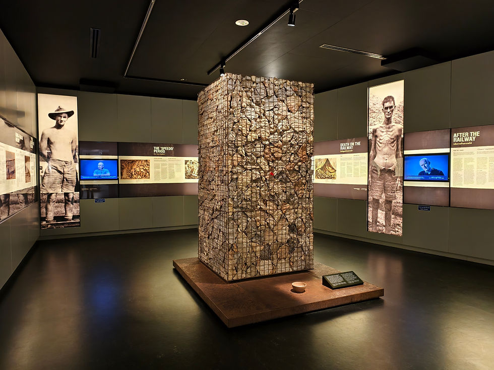 Museum exhibit of stone pillar and historical photos, dimly lit. Panels with text and two screens displaying a man. Mood is somber.