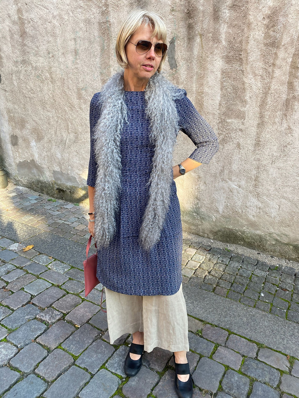 Woman in sunglasses wearing blue tdress, offwhite trousers and long lightgrey sheepskin shawl and holding red clutch