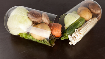 Two transparent capsules filled with vegetables like cabbage, potatoes, and mushrooms on a dark surface, symbolizing nutrition and health.