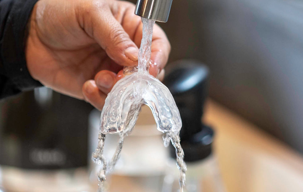 Hand rinsing a transparent dental mouthguard under running water from a silver tap, with a blurred kitchen background.