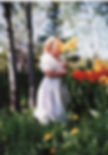 me in a white dress with puff sleeves around the age of two or three in the early 90s. i am smelling flowers. my face is in a bouquet of yellow daffodils with red and yellow tulips blooming behind me. a bed of luscious green grass and dandelions is in the foreground, and birch trees line the background. a banging photo which gives me life to this day. film photo taken by my talented mama Kris Monson.