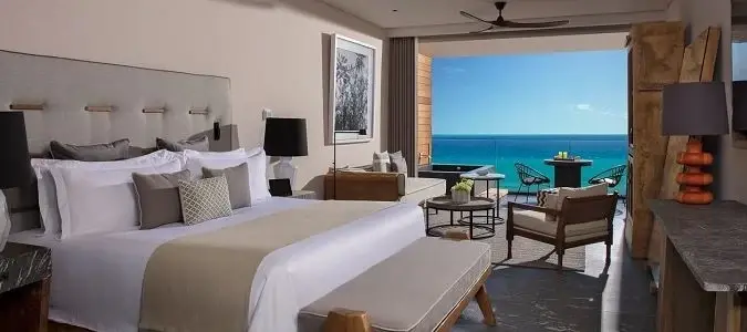 Modern seaside bedroom with king bed, beige accents, and balcony seating overlooking a bright blue ocean and sky