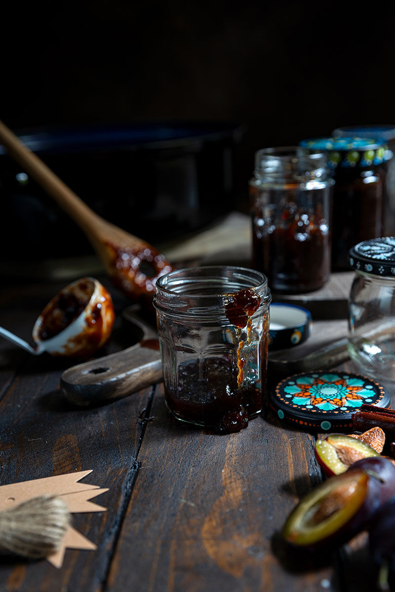 Moody still life of open jars of dark plum butter, powidl, wooden spoon, and sliced plums on a rustic table with colorful patterned lids.