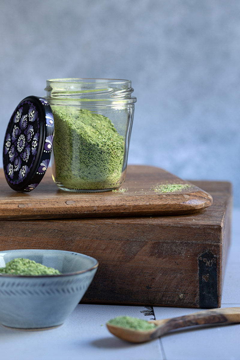 Glass jar of green wild garlic salt with mandala lid on a wooden box, beside a bowl and spoon on a blue background.