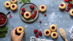 Overhead view of jam thumbprint cookies, Hallongrottor, with raspberries and currants on a blue table, with a hand reaching for one.