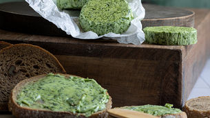 Sliced green wild garlic-butter on parchment and rustic bread on a wooden board, with a knife in a moody green-background kitchen.