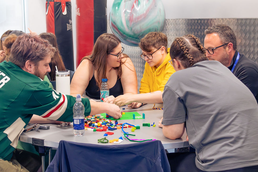 People gather around a table building with colourful LEGO bricks, chatting in a gym-like room with punching bags and a large exercise ball.