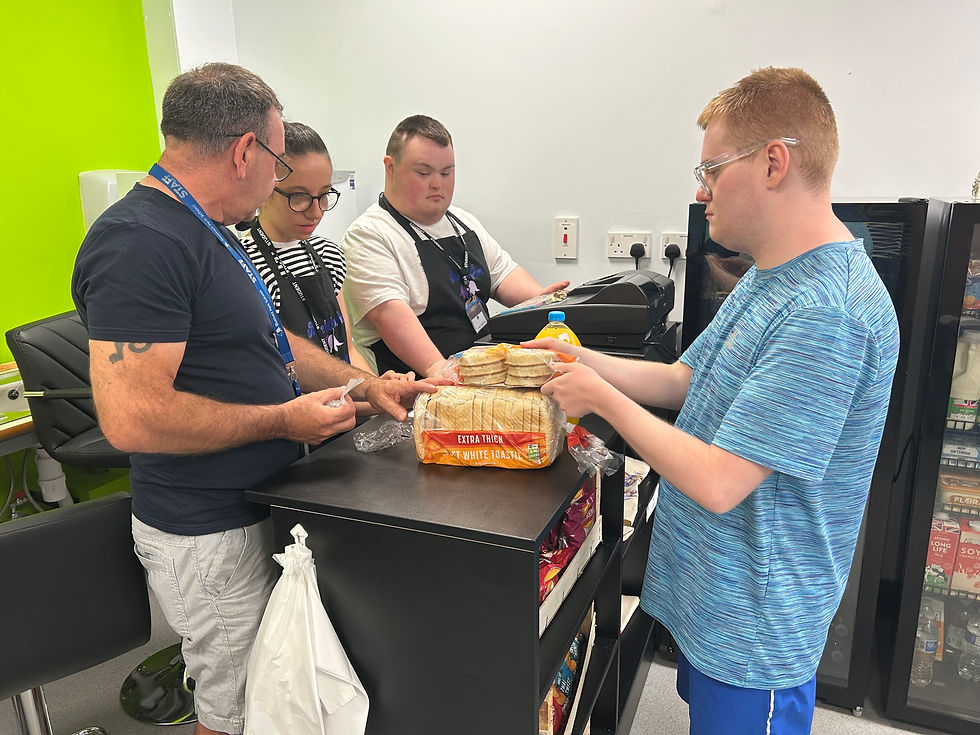 Four people in a small shop setting. Two students and a member of teaching staff are behind the counter and one young person is buying bread, crumpets and juice.