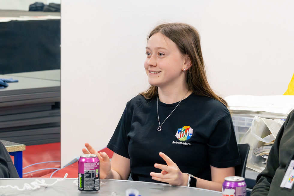 Smiling girl in a black Ambassadors T-shirt sits at a table with drinks cans in a classroom or workshop setting