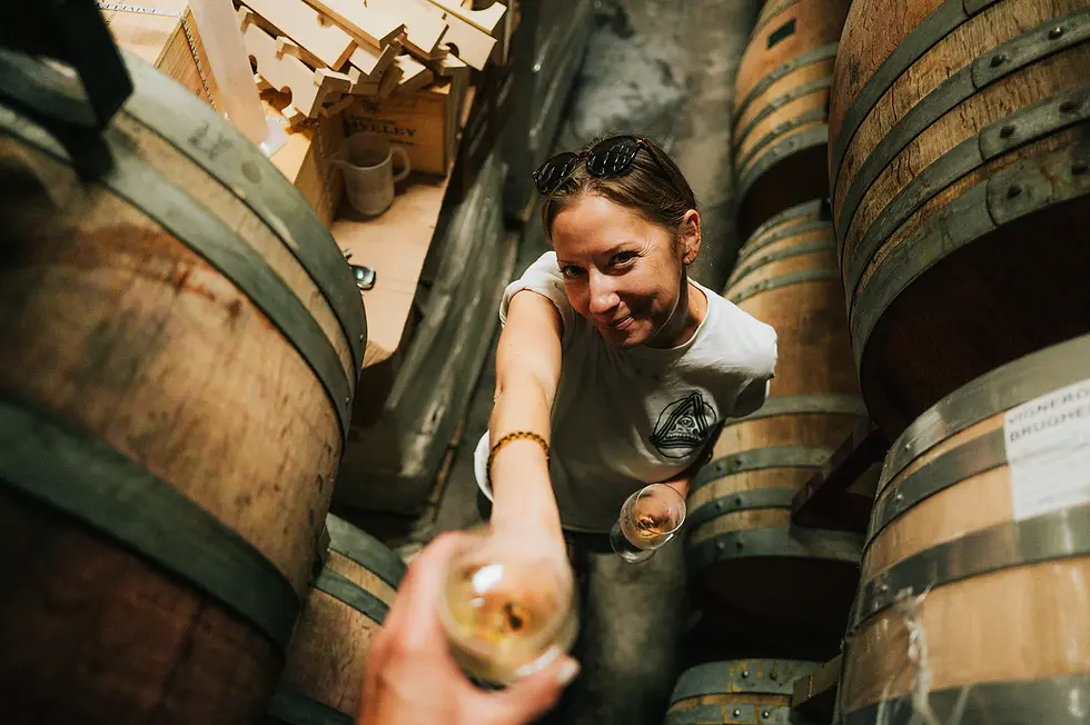 female winemaker with her barrels looking into the camera and giving a glass of wine to the viewer