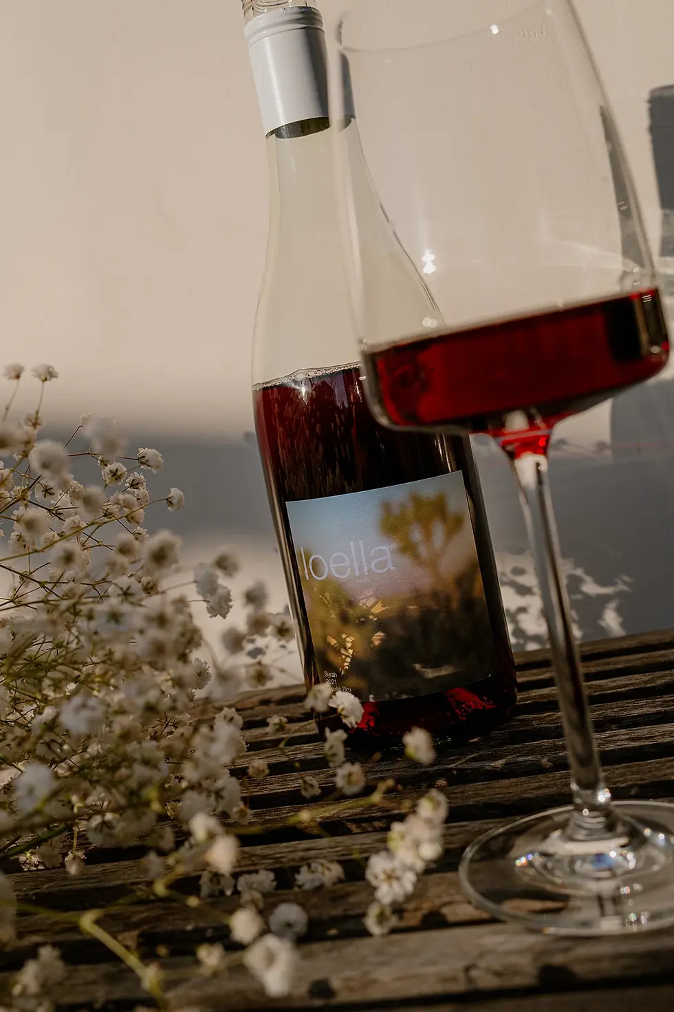 A bottle of red wine, Syrah is photographed on a sunny afternoon on the porch with flowers in the foreground