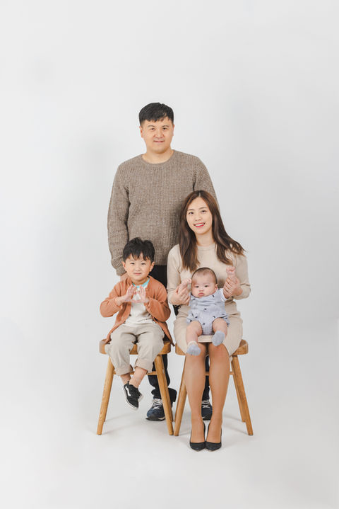 Studio portrait with eldest child seated left, mom holding baby on right, and father standing behind looking into the camera (왼쪽엔 첫째, 오른쪽엔 엄마와 아기, 뒤에는 카메라를 응시하는 아빠가 서 있는 가족사진)