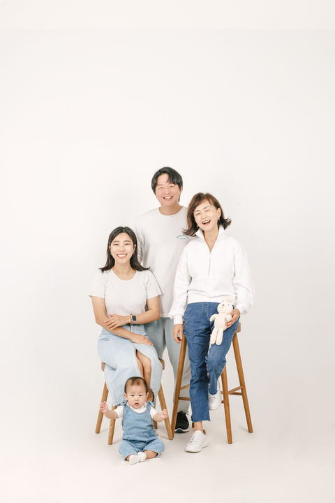 Three-generation family portrait – grandmother and mother seated, father standing behind, child sitting in front on the floor.
세대를 아우르는 가족사진 – 할머니와 엄마는 의자에 앉아 있고, 아빠는 뒤에 서 있으며, 아기는 앞에 바닥에 앉아 있음