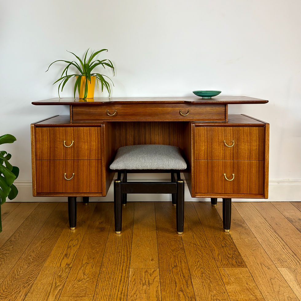 Mid-century wooden desk with drawers, gray stool, potted plant, and green bowl against a white wall on a hardwood floor.