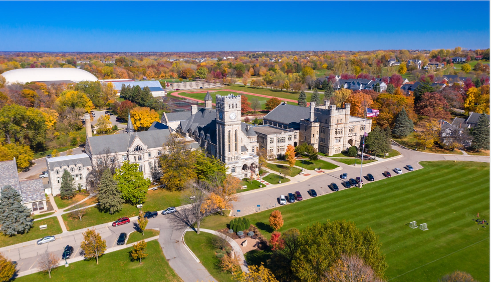 Aerial view of a stone campus building with a clock tower beside a green field and track, surrounded by colorful autumn trees.