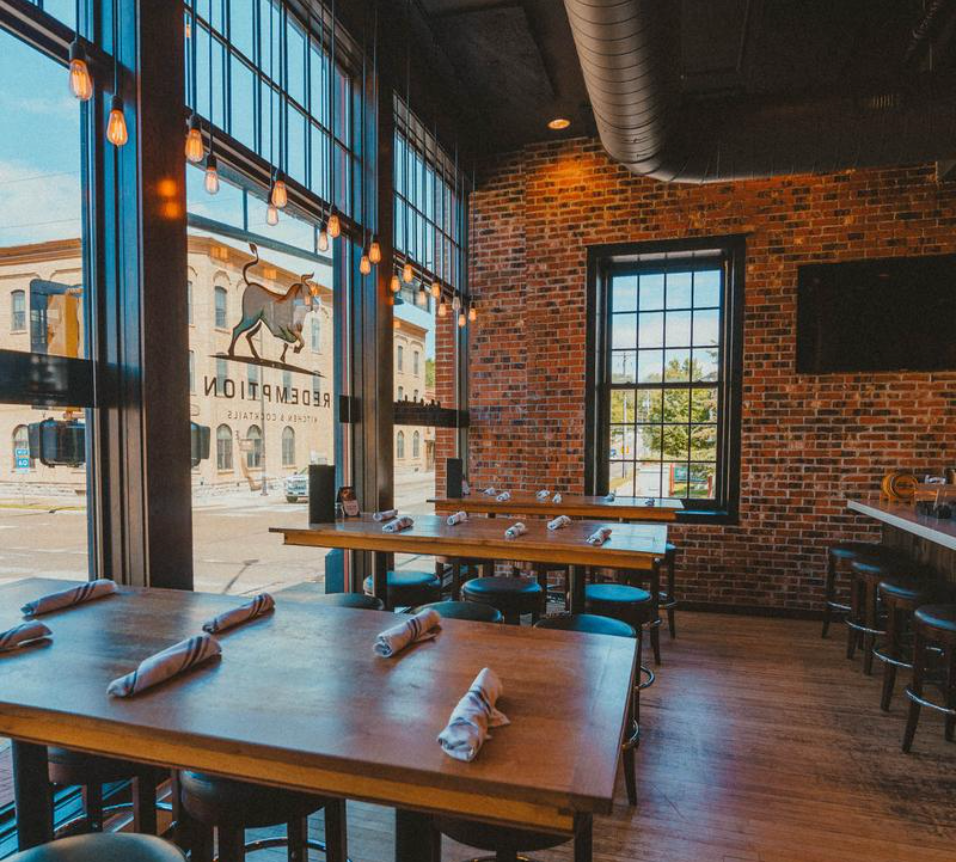 Brick-walled restaurant with large windows, set tables, and a reversed Redemption logo on the glass.