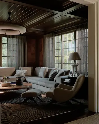 Cozy room with a gray sectional sofa, armchair, and footstool on a patterned rug. Large windows with sheer curtains, a lamp on a side table.