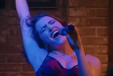 Hannah singing into a microphone expressively. She is lit with blue light and standing in front of a brick wall. Still from The Deal on Amazon Prime.
