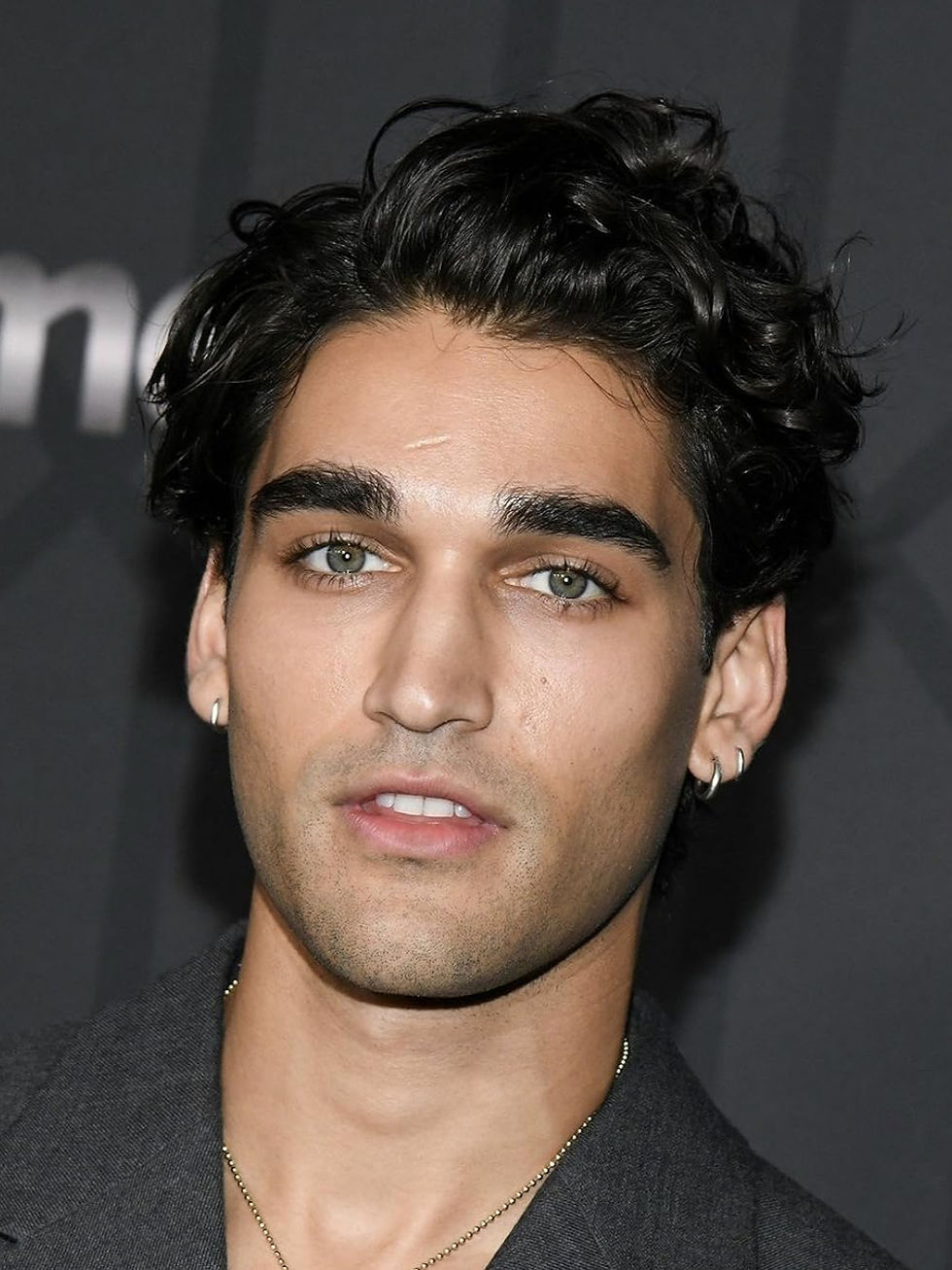 A picture of Off Campus actor John Heuston. He has dark curly hair and light green eyes wears a dark jacket and earrings. He has a neutral expression.