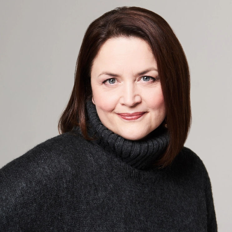 Ruth Jones headshot. She wears a black poloneck jumper and has dark brown hair and green eyes. She is looking directly at the camera and smiling slightly.