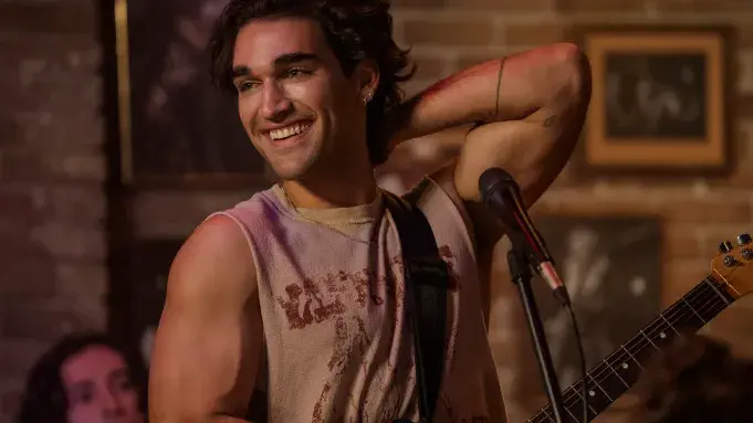 Justin is smiling with his guitar beside a microphone on a small stage, brick wall and framed art behind him. Still from The Deal on Amazon Prime