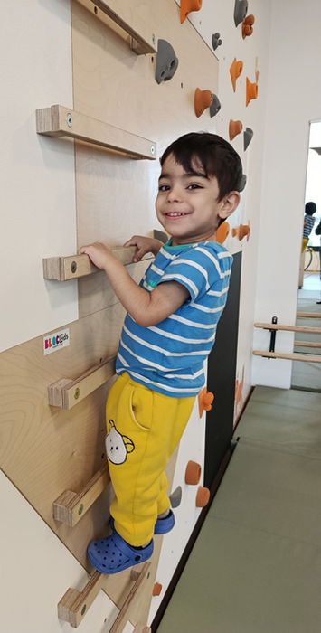 Ein Kind klettert mit einem Lächeln an einer kindgerechten Kletterwand im Bewegungsraum. Die Aktivität fördert Motorik, Koordination, Gleichgewicht und stärkt das Selbstvertrauen durch spielerische Bewegung.