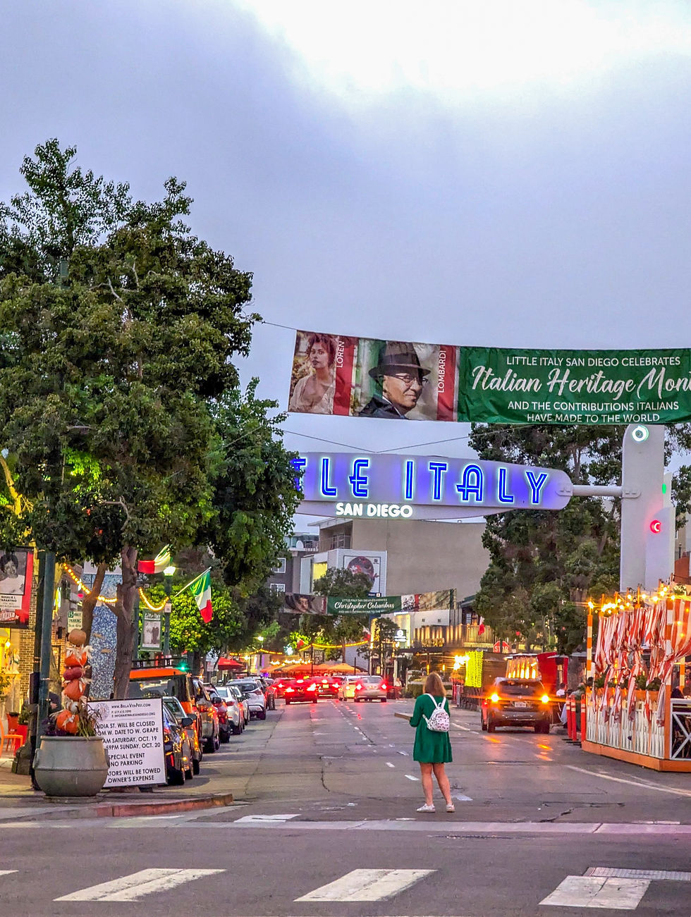 Woman in green dress stands in middle of busy street under a neon blue sign that reads, "Little Italy San Diego"