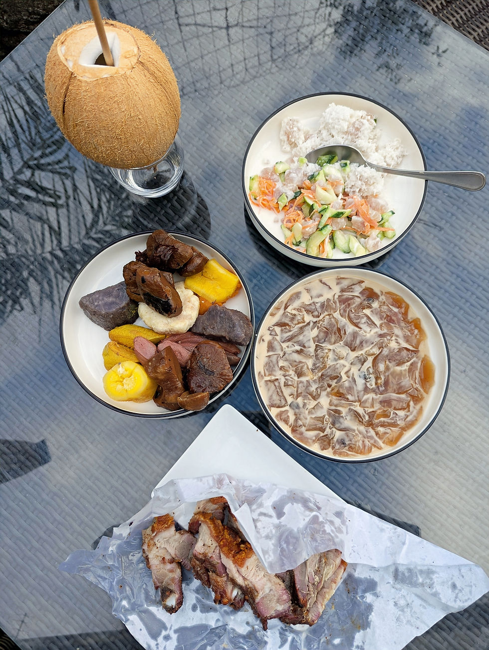 A table display of 5 traditional dishes of Tahiti: Fresh coconut water, poisson cru, a tapioca-like dessert, roasted pork, and a plate of taro root, breadfruit and bananas.