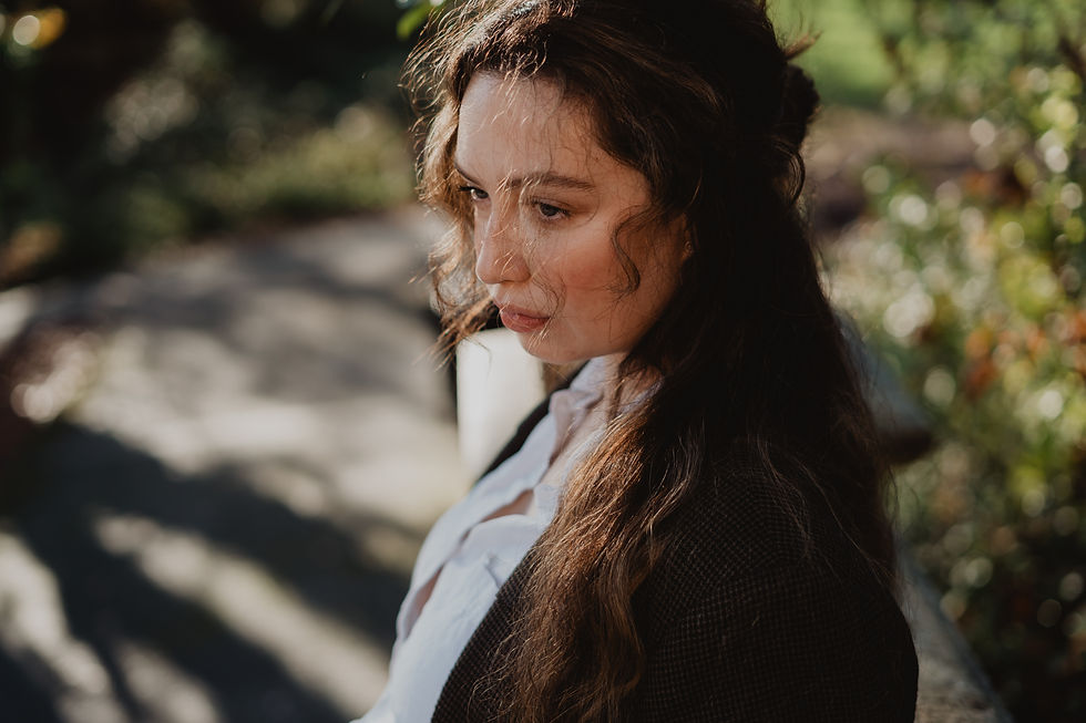 Woman with long hair gazes pensively outdoors, wearing a dark jacket and white shirt. Sunlight creates soft contrasts and a serene mood. - Jane Austen inspired bookish photoshoot at Royal Holloway