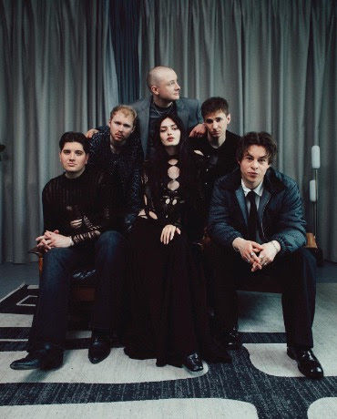 Five people in dark clothes pose on a couch before gray curtains, wearing serious expressions in a moody studio scene.
