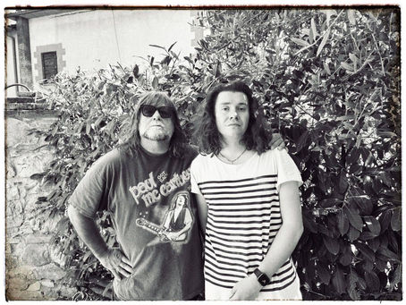Two men pose in black-and-white by leafy bushes; one wears sunglasses and a Paul McCartney T-shirt, the other a striped shi