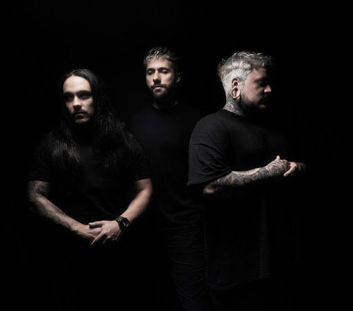 Three tattooed men in black clothing stand against a dark studio background, lit dramatically with a moody serious look.