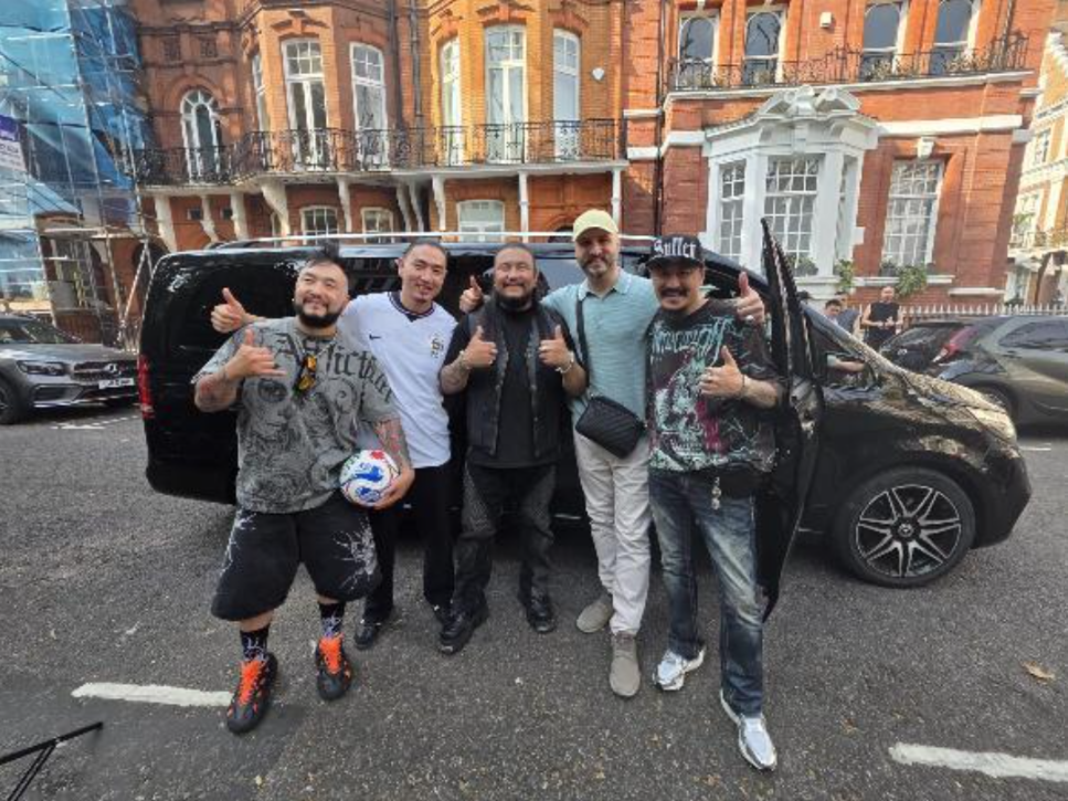 Five men pose with thumbs up beside a black van on a city street, in front of a red-brick building, smiling and upbeat.