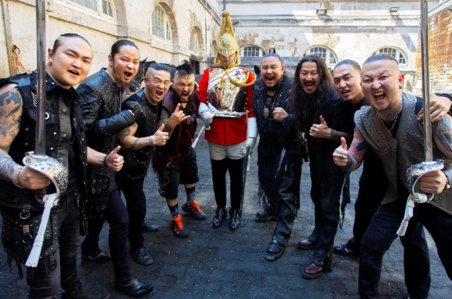 Group of men in black pose and shout around a ceremonial guard in a red uniform, holding swords in a courtyard.